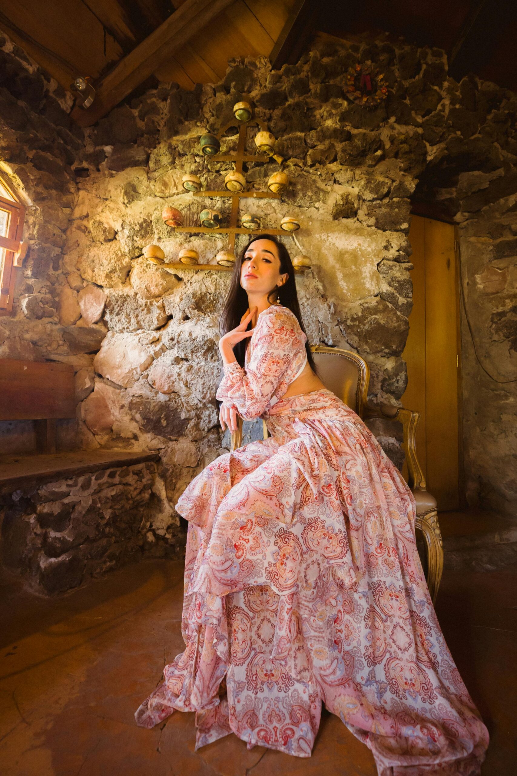 A woman in a floral gown poses gracefully inside a rustic stone cottage with warm lighting.