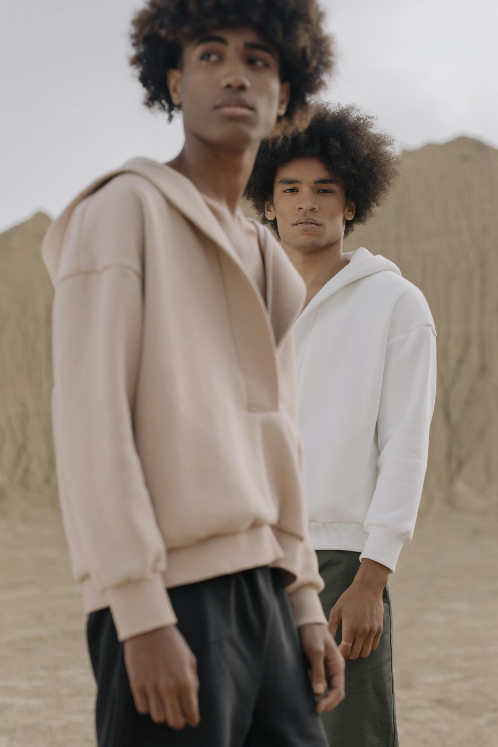 Two stylish young men in hoodies stand in a serene desert landscape, showcasing modern fashion.